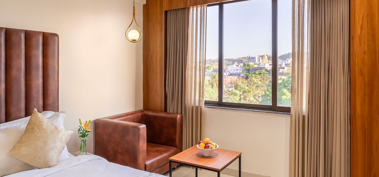 A cozy hotel room includes a comfortable bed, a brown armchair, a small table with fruit, and a window with a city view - Lords Eco Inn Ambaji.