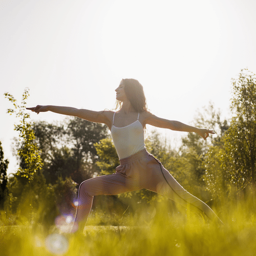 Woman practicing yoga outdoors in a grassy field, holding Warrior II pose with arms extended, surrounded by sunlight and trees