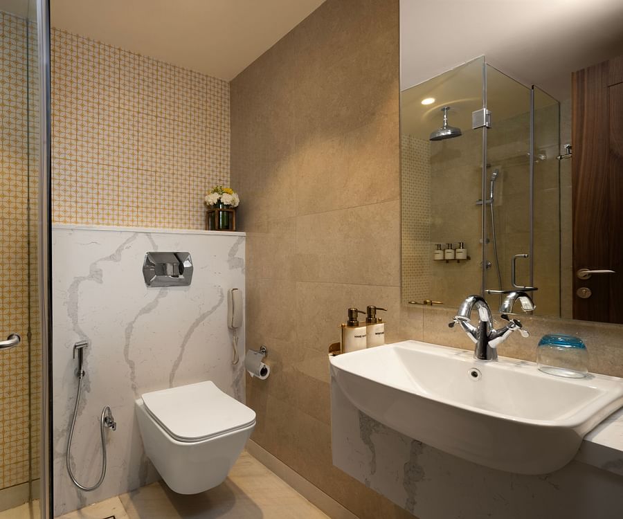 A sleek, modern bathroom with a glass-enclosed shower, beige and white patterned tiles, a marble-look countertop with a white sink, chrome fixtures, wall-mounted toiletries, and a touch of freshness from a vase of yellow and white flowers - Aloha On the Ga