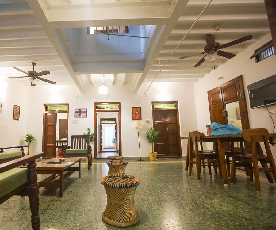 alt-text A wide area of the living room with wooden furnished table , sofa, stool at A top view of the room with plushy bed and the wall painting at la villas Heritage, pondicherry