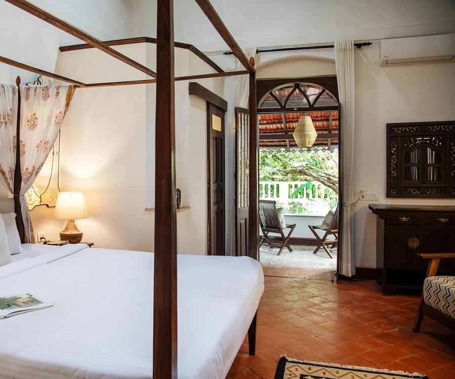 An image of a bed with four posters, a bedside table with lamp, a wardrobe, and open wooden door at Lamrin Ucassaim Hotel, Goa