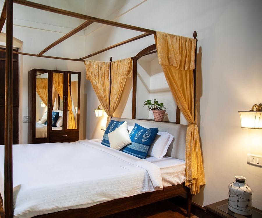 An image of a four poster bed, bedside table, wooden dressing table, and other wooden furnishings at Lamrin Ucassaim Hotel, Goa