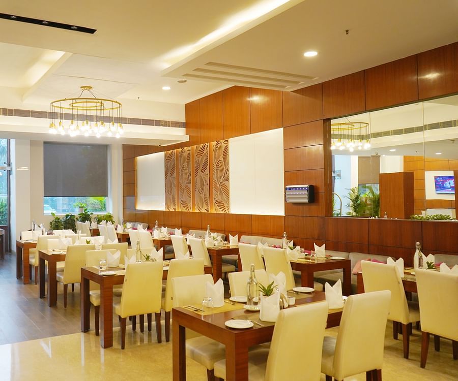 Spacious and elegant dining room at Nemesia with multiple tables set with white tablecloths and chairs. The room features large windows, wooden paneling, and a chandelier, creating a warm and inviting atmosphere.