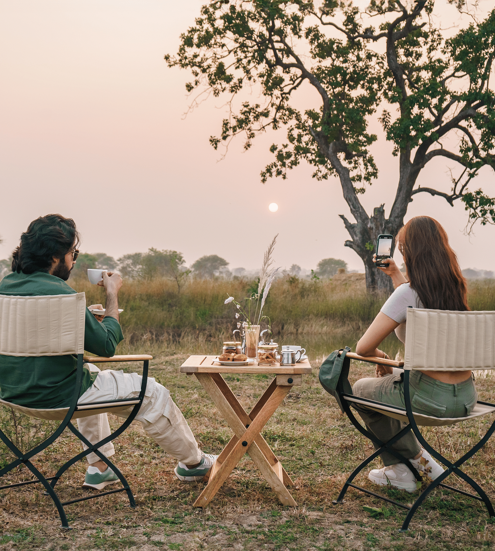 Two people sitting on chairs with a dining setup in an open grassland surrounded by trees at Trees N Tigers, Tadoba.