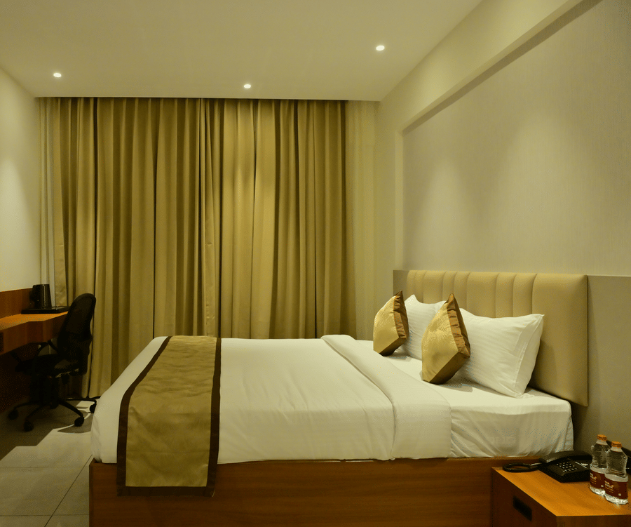 Deluxe room at Bhavanis Vista By Icon Hosur with a king bed, warm drape curtains, and a wooden TV console.