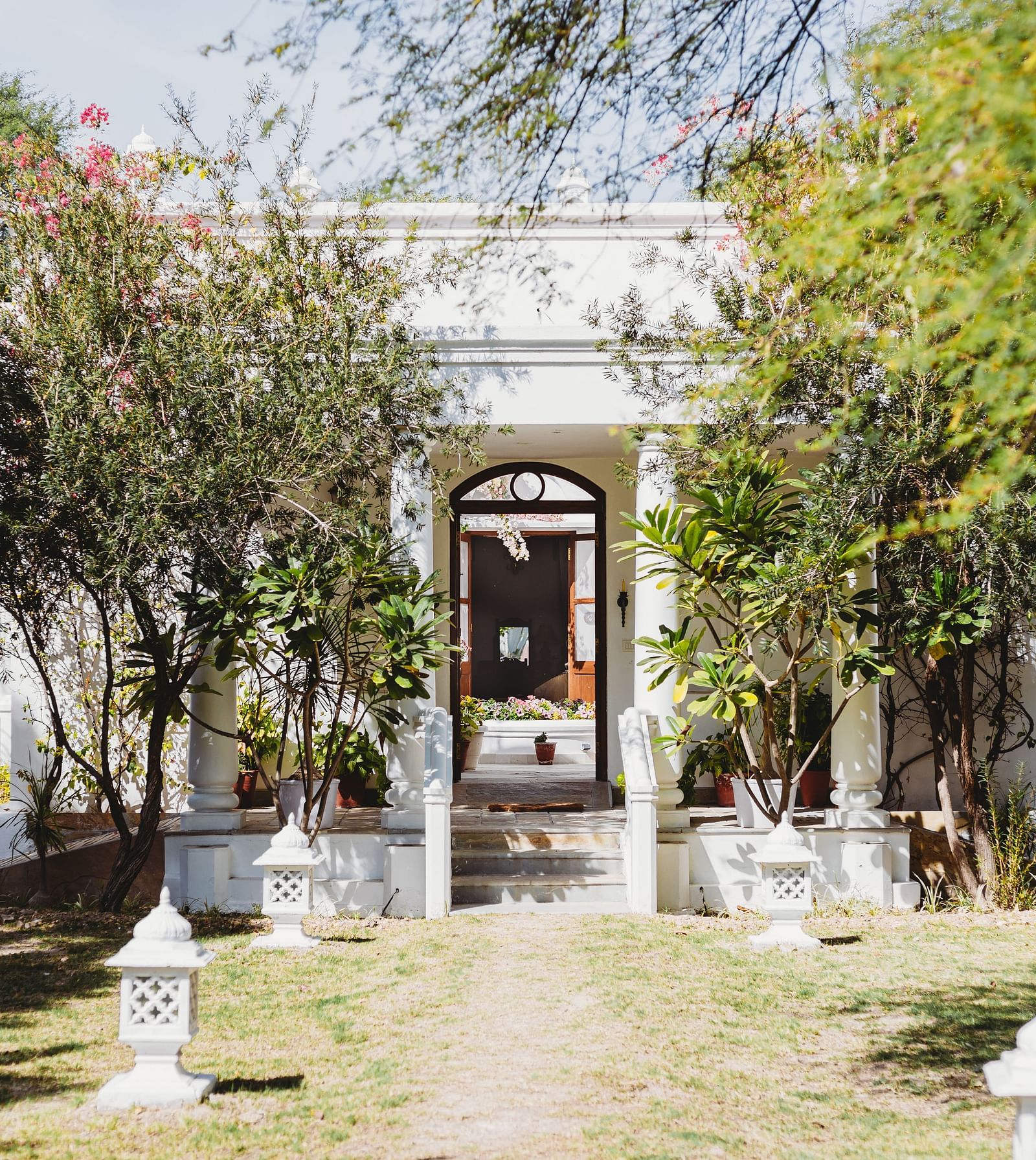 A white building with an arched doorway is framed by greenery and trees, with a pathway leading to the entrance- Shahpura Bagh.