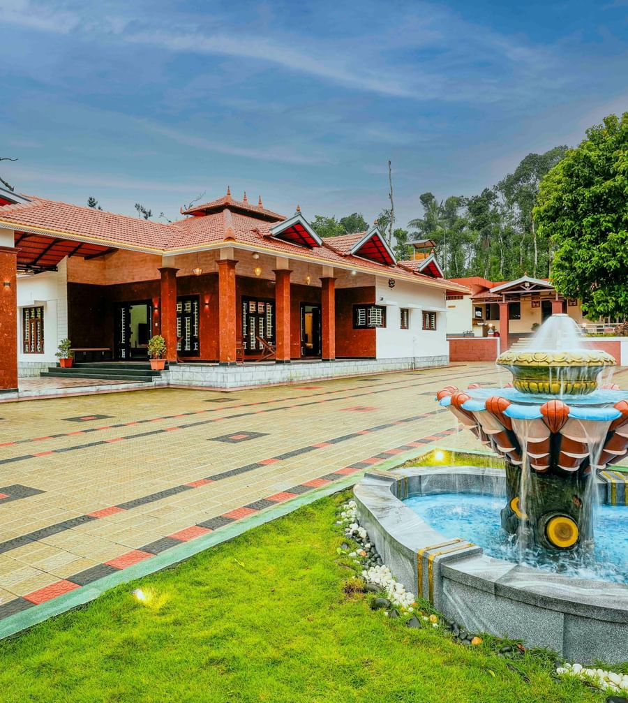 A wide-angle view of the central courtyard at Bhuthanakadu Retreat, featuring a decorative fountain and the traditional architecture of the buildings under a bright, cloudy sky.