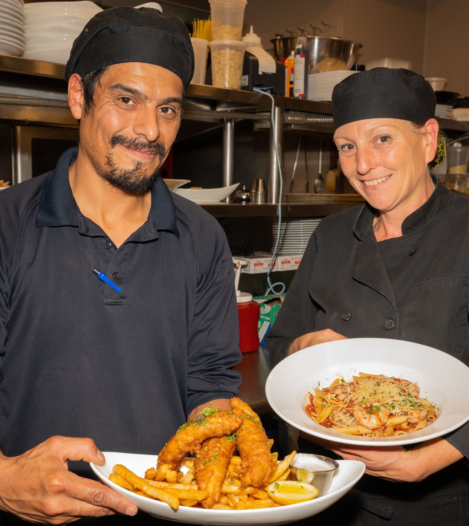 Two chefs smiling while holding plated dishes in the kitchen at The Groveland Hotel.