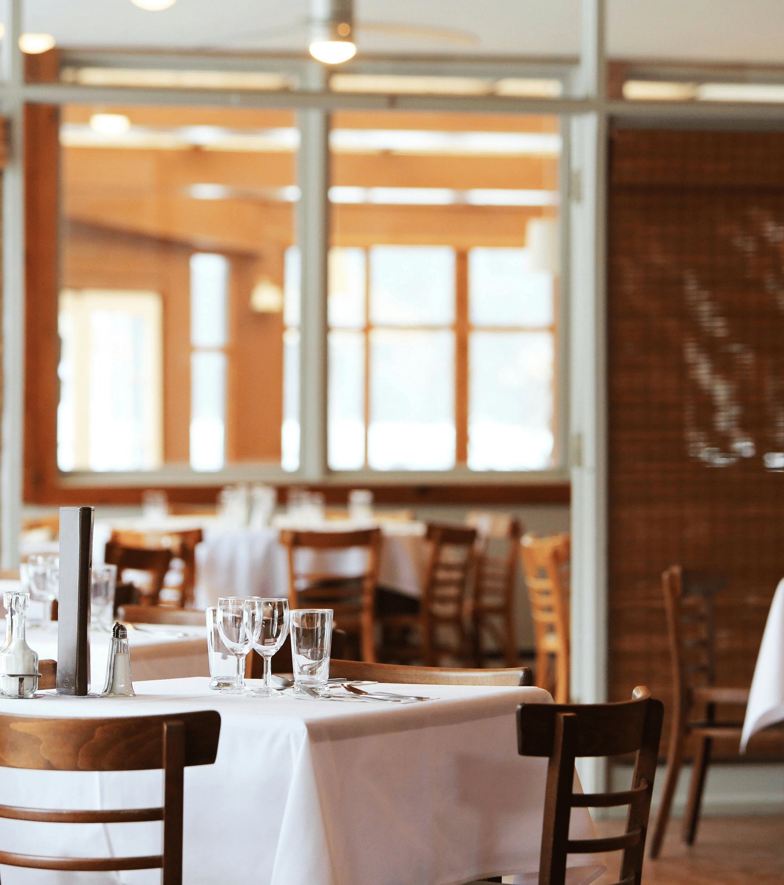 The bright interior of a restaurant with wooden tables and chairs, crisp white tablecloths, and bamboo window blinds.