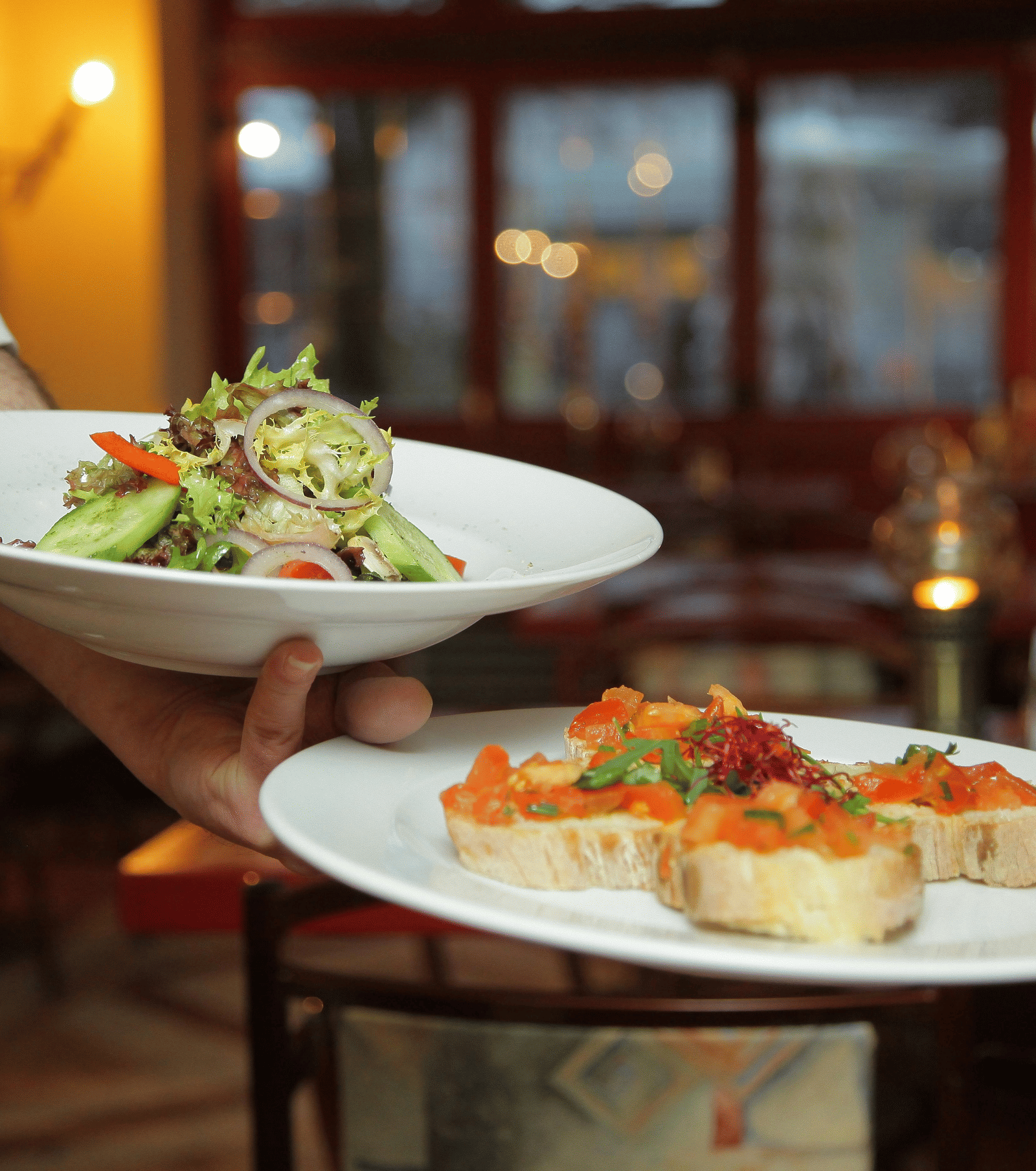 A waiter in a white shirt and black waistcoat is serving two plates of food, one with a side salad and one with bruschetta.