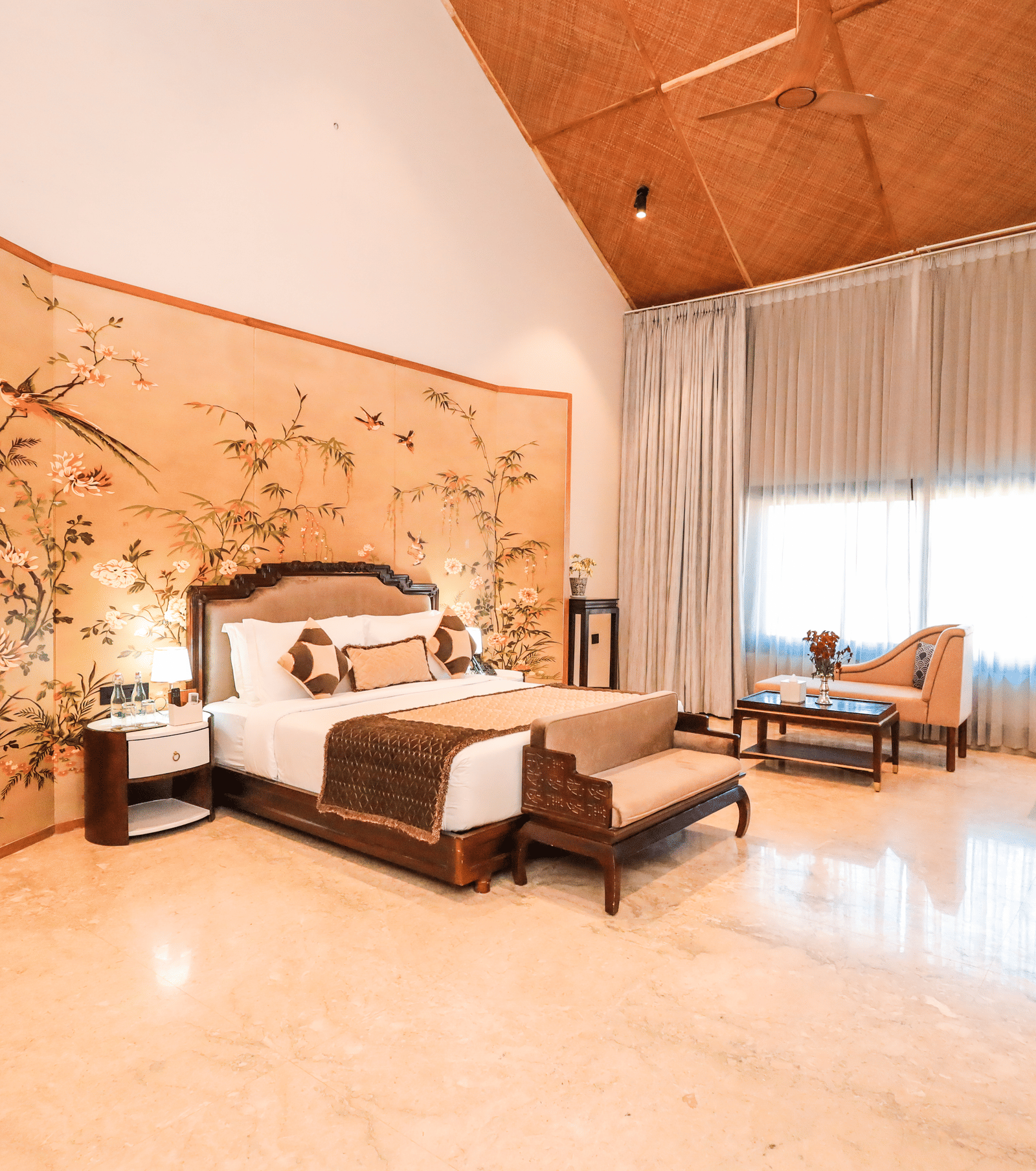 A room shows a bed with side tables, a bench at the foot of the bed, a seating area with chairs and a large window with curtains at Ananta Spa & Resort, Jaipur.