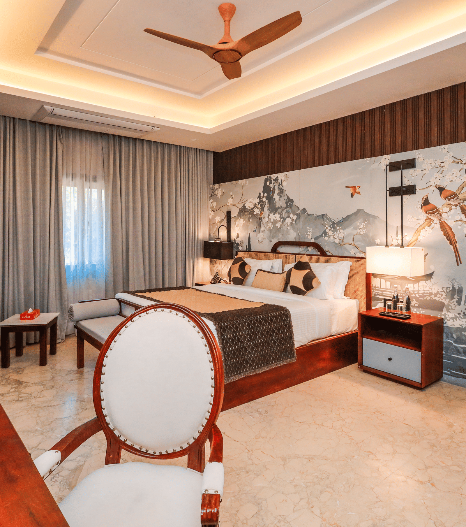 A room shows a bed with side tables, chairs with a table, curtains on windows and storage units along a wall at Ananta Spa & Resort, Jaipur.