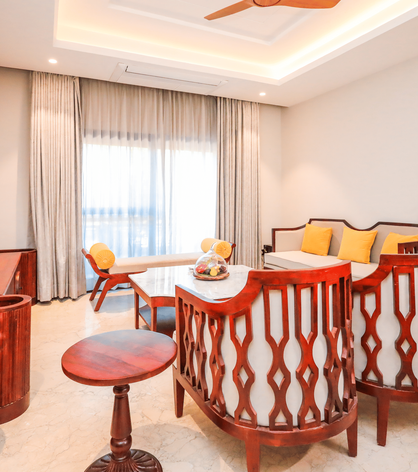 A room shows a table with stools, a long seating area with cushions, a window with curtains and a door to another space at Ananta Spa & Resort, Jaipur.