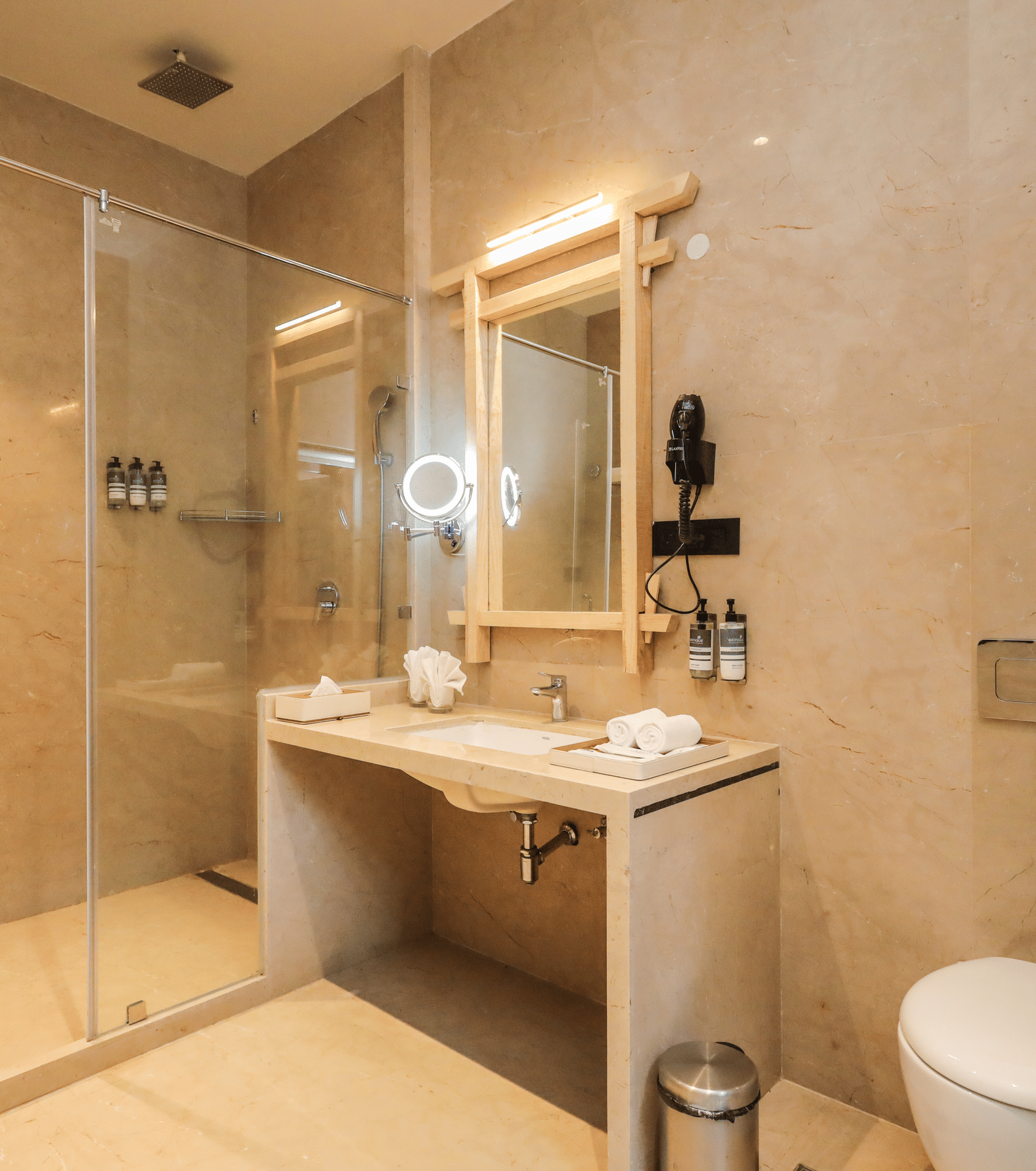 A washroom shows a basin with a mirror, a glass shower enclosure, a toilet and toiletries placed on the counter at Ananta Spa & Resort, Jaipur.