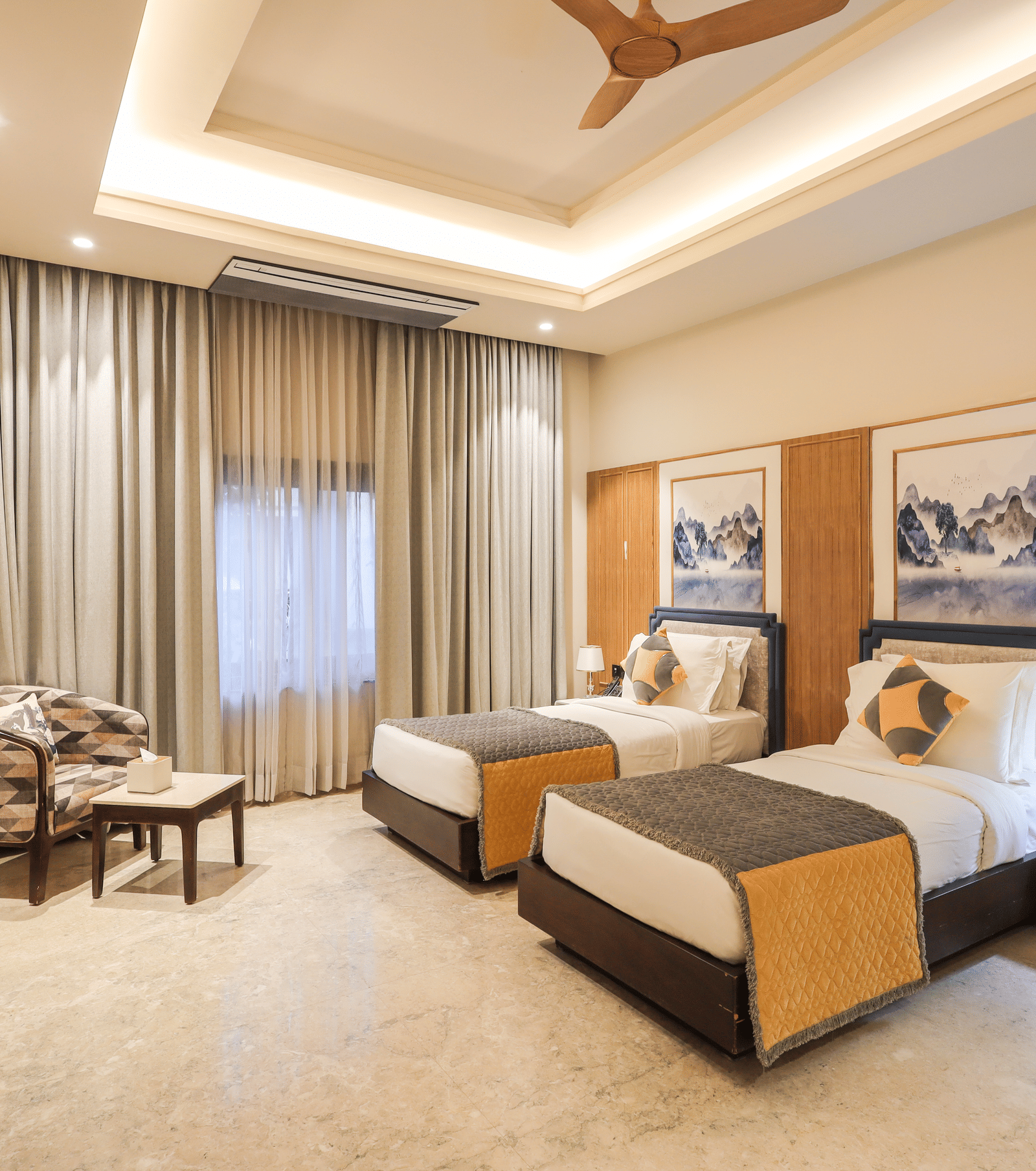 A room shows two beds with side tables, a seating table with chairs, a window with curtains and overhead lighting at Ananta Spa & Resort, Jaipur.