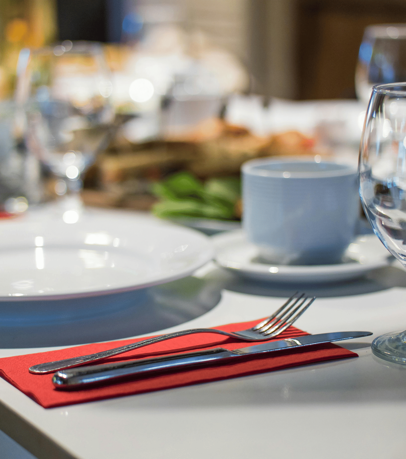 A close-up, blurred shot of a formal dinner table setting with white plates, red napkins, silverware, and clear wine glasses.