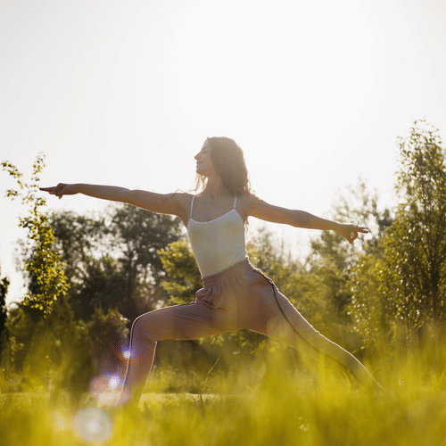 Woman practicing yoga outdoors in a grassy field, holding Warrior II pose with arms extended, surrounded by sunlight and trees