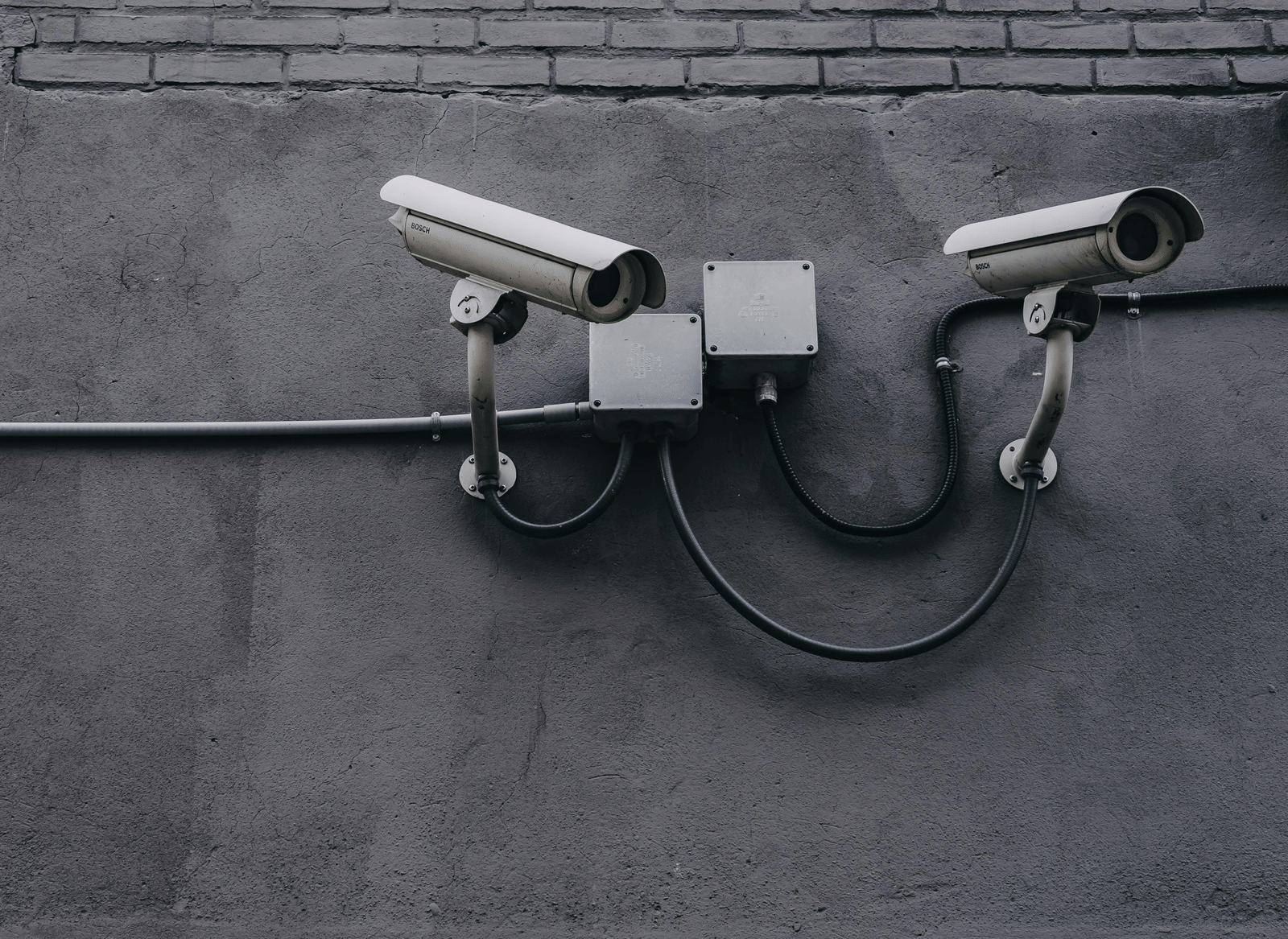Two surveillance cameras mounted on a grey concrete or brick wall, with visible wiring.