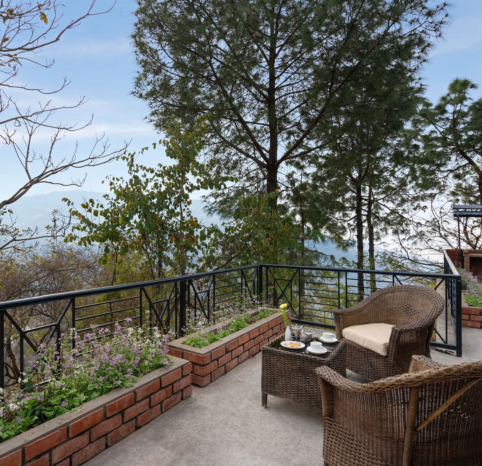 Two wooden chairs and a table set on an open balcony with a view of trees and mountains afar - Baikunth resort, kasauli 