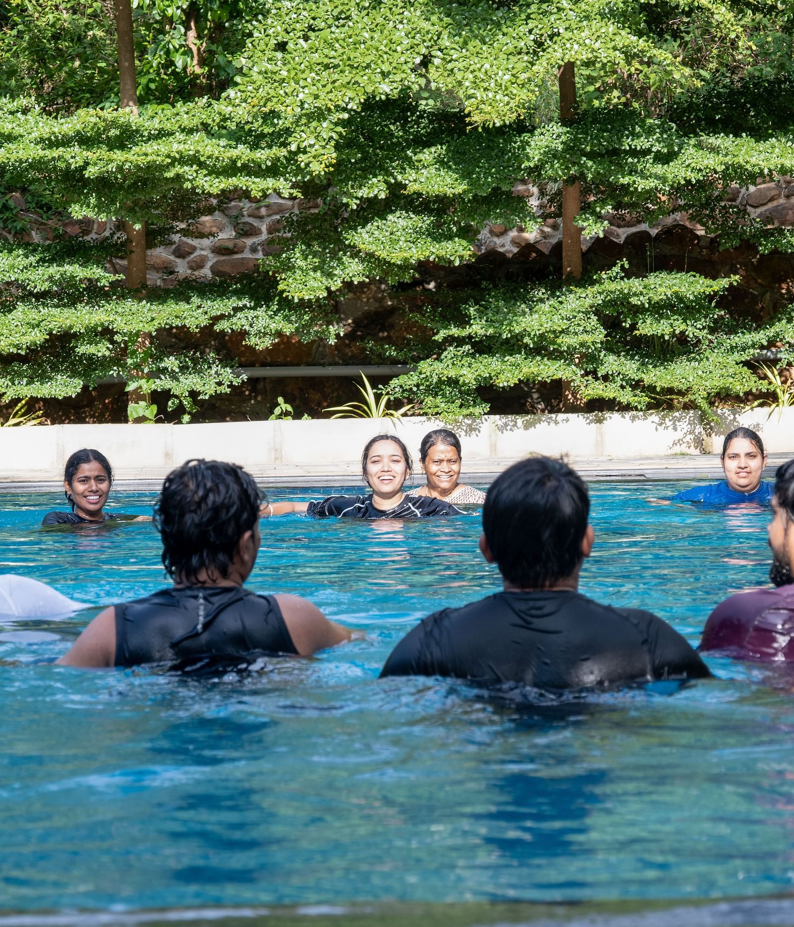 A close-up view shows multiple individuals submerged and swimming in a blue pool, with the background filled with green trees | RD's Nature Retreat, Bangalore