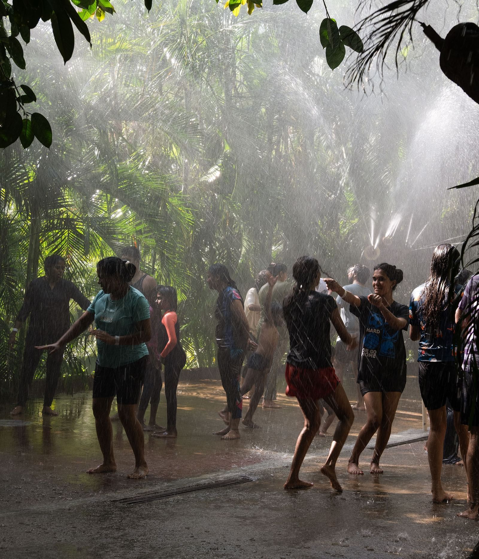 A group of people are seen enjoying a rain dance or water activity, with water spraying down on them in a shaded, outdoor setting | RD's Nature Retreat, Bangalore