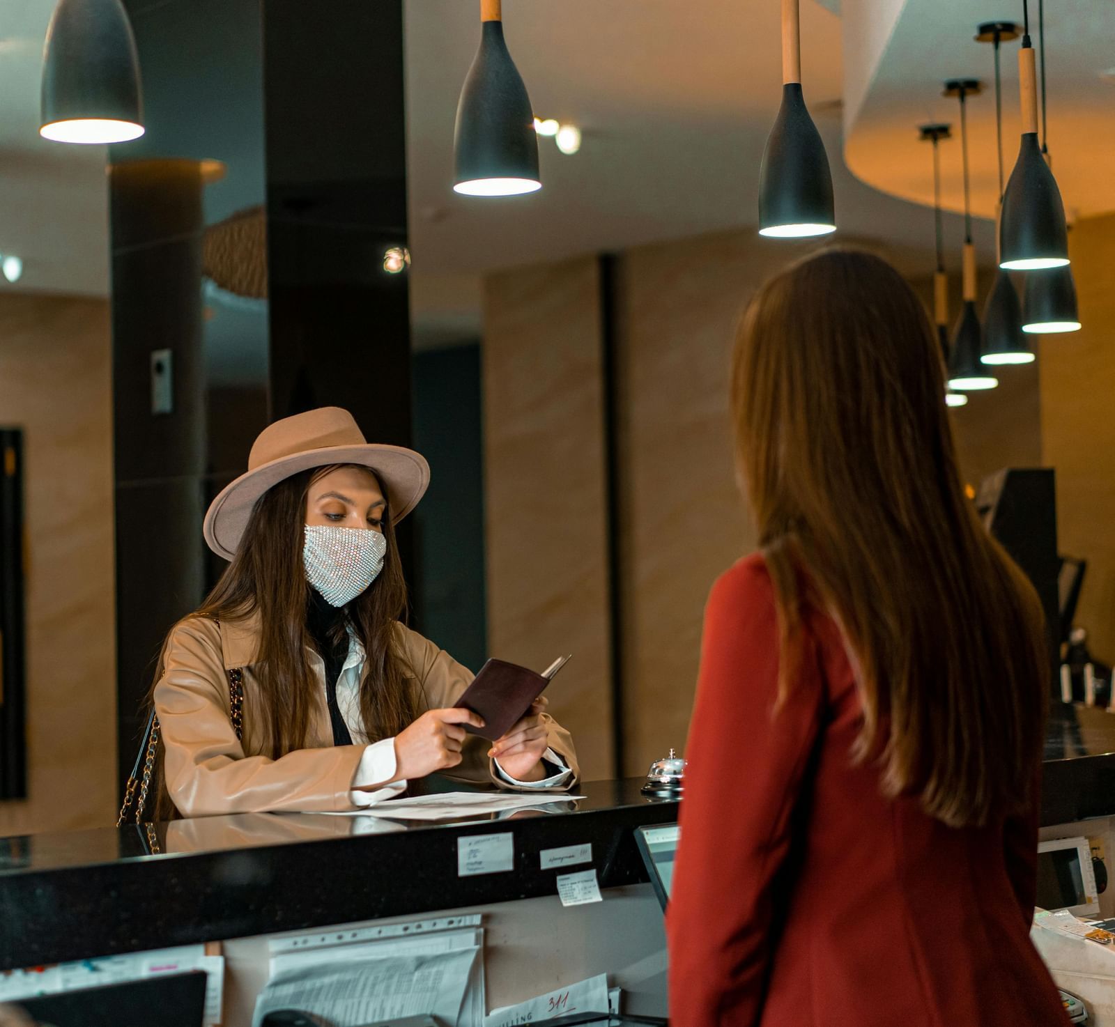 A guest wearing a face mask and a hat with a smartphone in her hand talking to the receptionist.