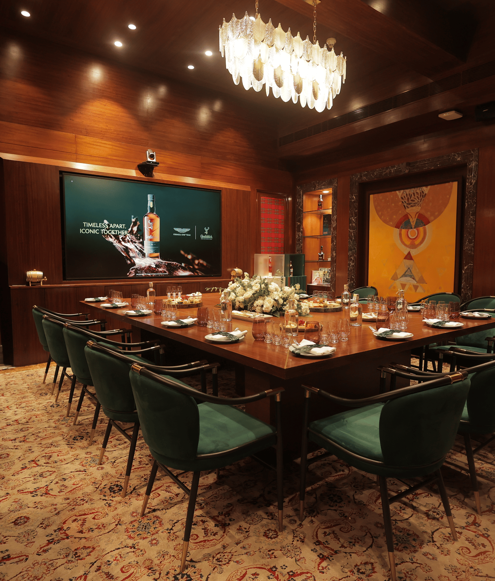 Wide view of an elegant private dining room featuring a large wooden table surrounded by green velvet chairs and a crystal chandelier at The Claridges New Delhi.