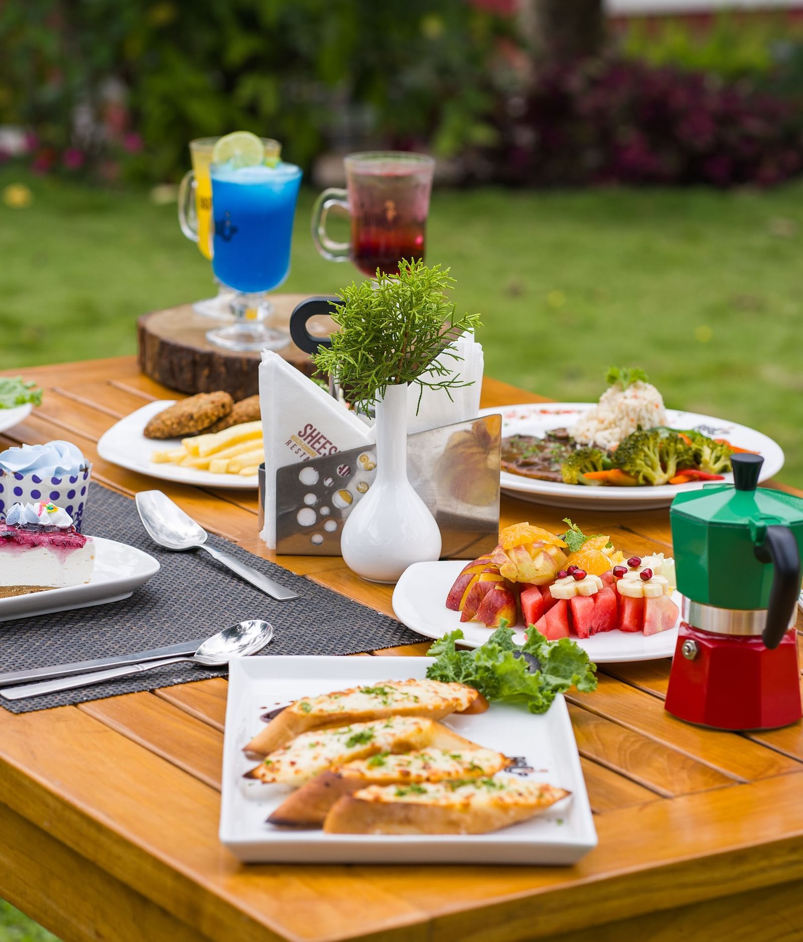 A wooden table is set outdoors with a variety of dishes and drinks, surrounded by a green lawn | Woodstock Resorts, Coorg