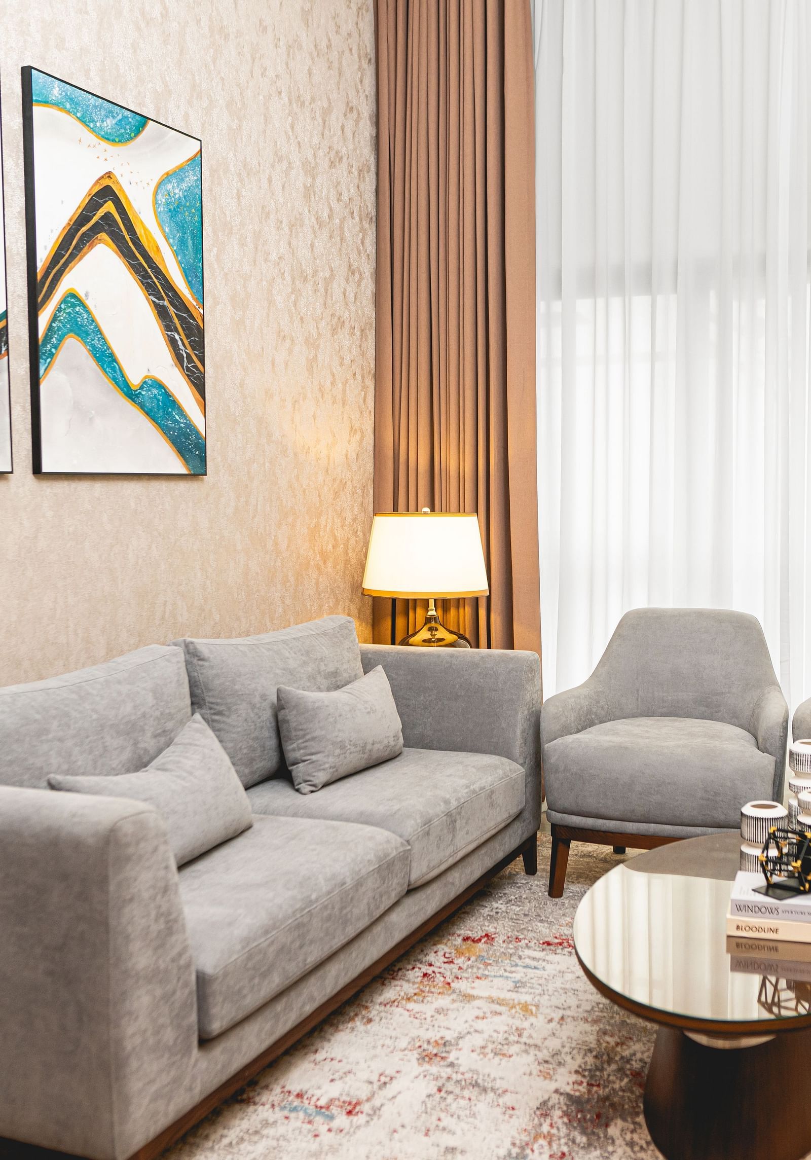 Room at KertelSuites with a sofa, two matching armchairs, a round coffee table, two floor lamps, a patterned rug, and abstract art on the wall. Large windows with sheer curtains allow natural light in.