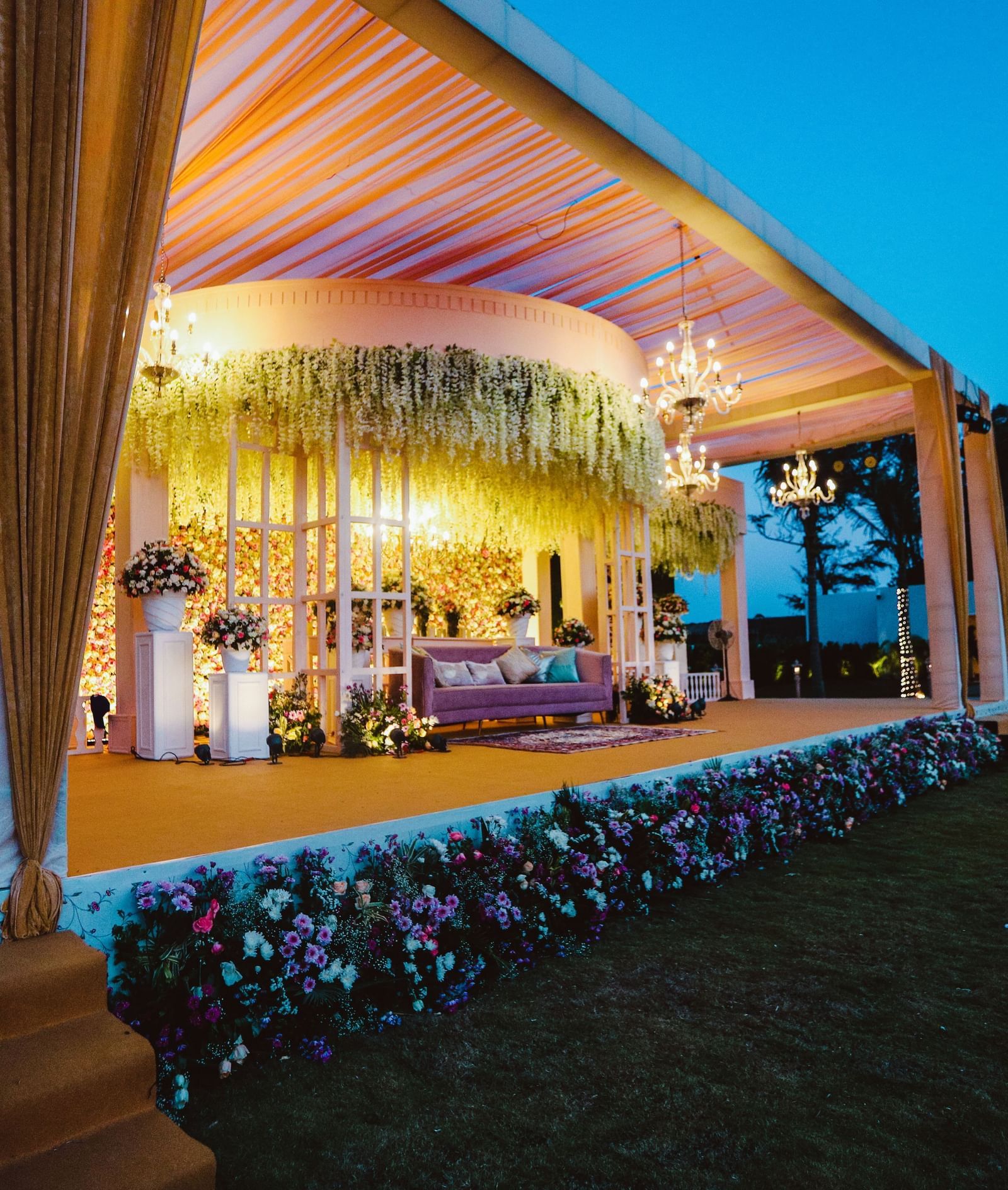 A stage decorated with flowers at the front set in a lawn, featuring ornamental plants, a chic sofa and chandeliers hanging from ceiling - Kaldan Samudhra Palace