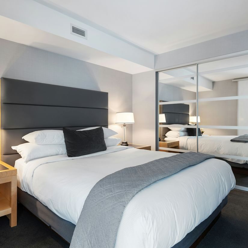 A modern hotel room with a double bed, a table with a lamp beside it and a huge mirror.