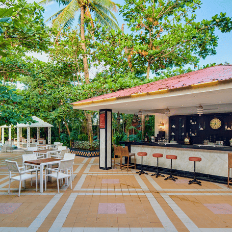 A pool bar area outside at MAYFAIR Palm Beach Resort, Gopalpur-on-Sea. Stools stand along a counter under a roof structure. Tables and chairs occupy a patio. Trees and plants surround the location.