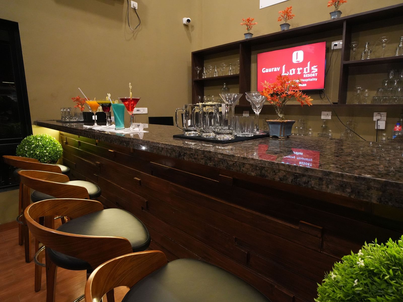 Close-up view of a modern bar setup with wooden high chairs, granite countertop, glassware, vibrant cocktails, decorative plants, and neatly arranged shelves in the background - Gaurav Lords Resort Shrivardhan