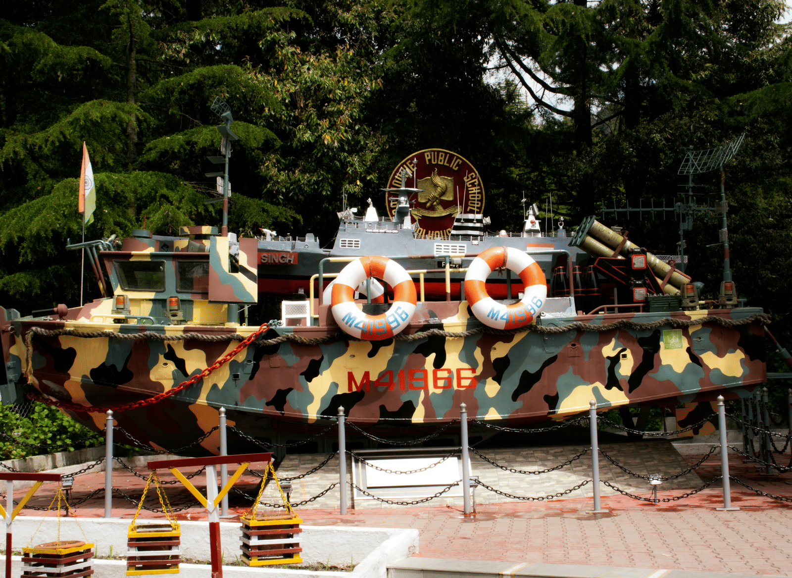 A camouflaged boat display with 2 lifebuoys and an emblem in the back, surrounded by trees and fencing.