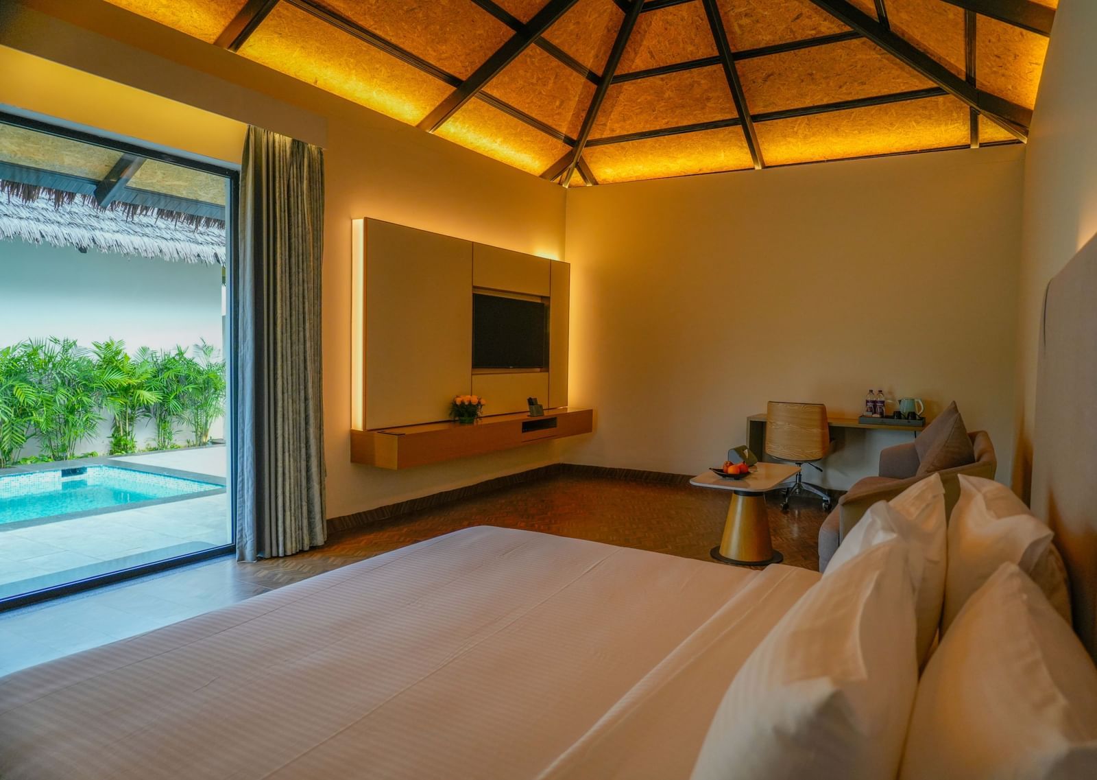 Interior view of a luxury villa room with a neatly made bed, warm ambient lighting, a mounted TV, a sitting area with a desk and chair, and a view of a private pool through the open sliding glass door - The Shore Trishvam, Pondicherry