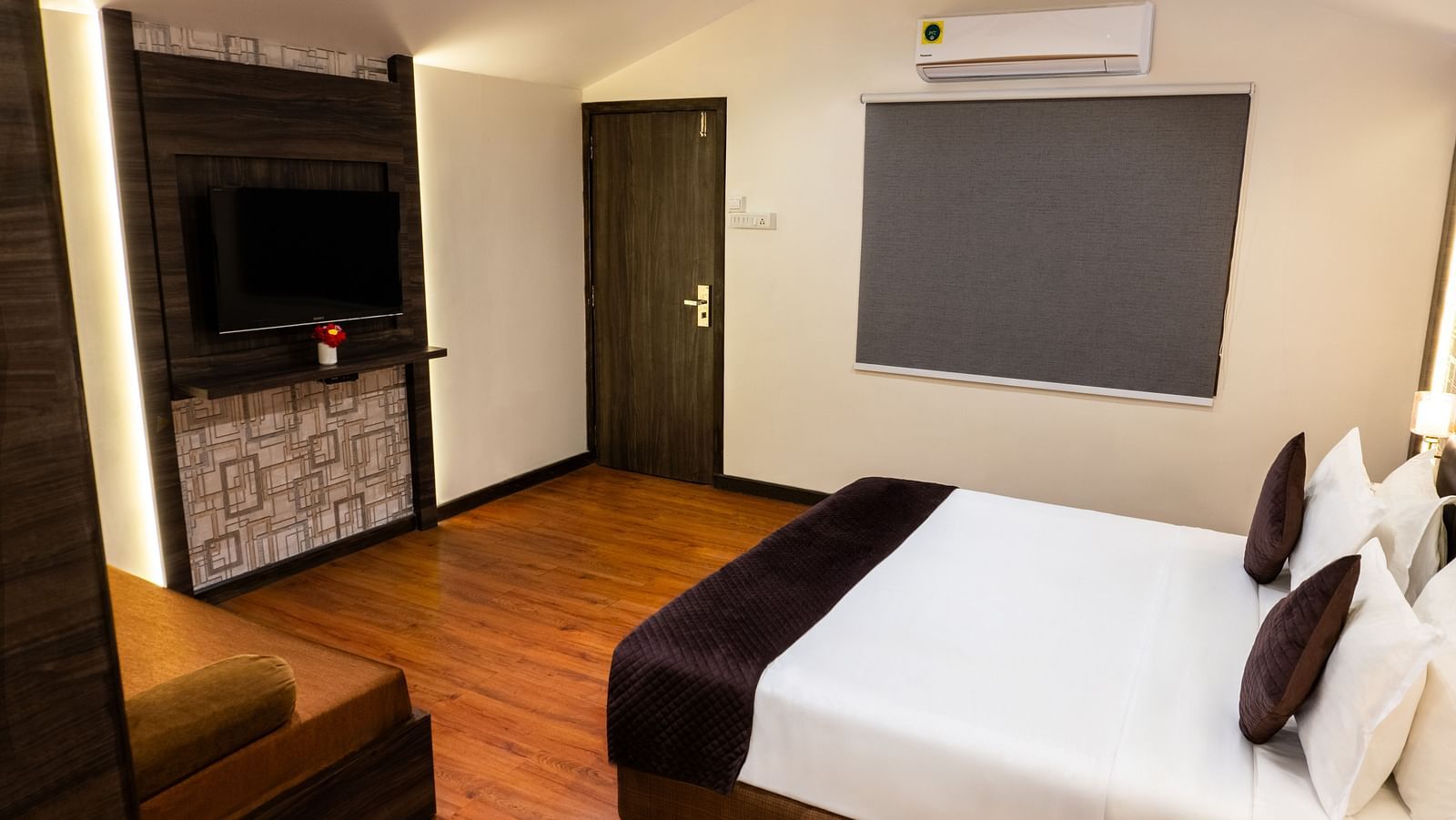 Hotel room interior featuring a bed with dark brown and white bedding, a flat-screen TV mounted on a wooden panel, wooden flooring, and an air conditioner above a covered window - The Grand Legacy Resort & Spa