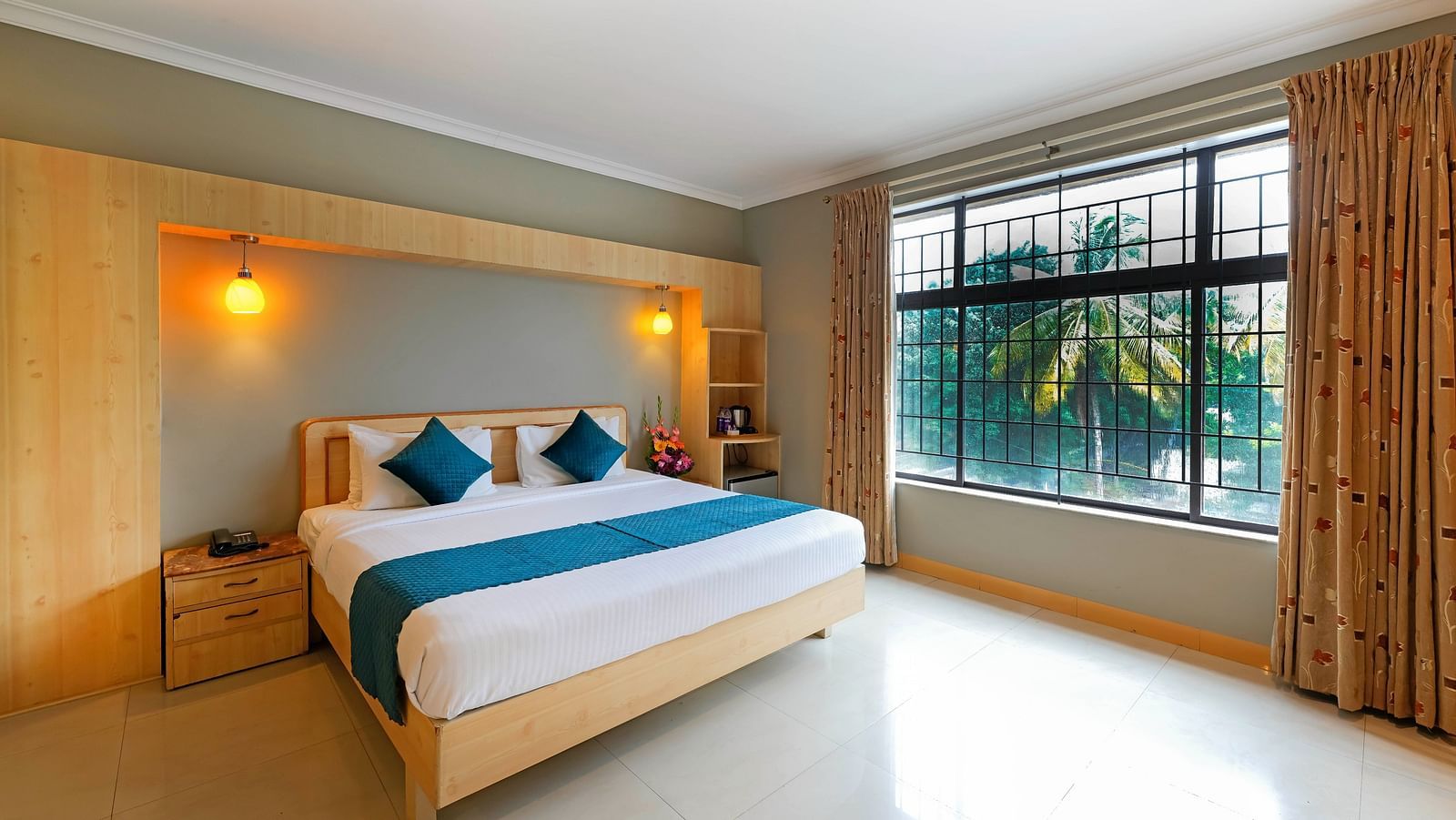 An image of a hotel room in Bengaluru featuring a queen size bed, a night stand, and sunlight from the expansive windows - Cross Roads Inn, Bengaluru