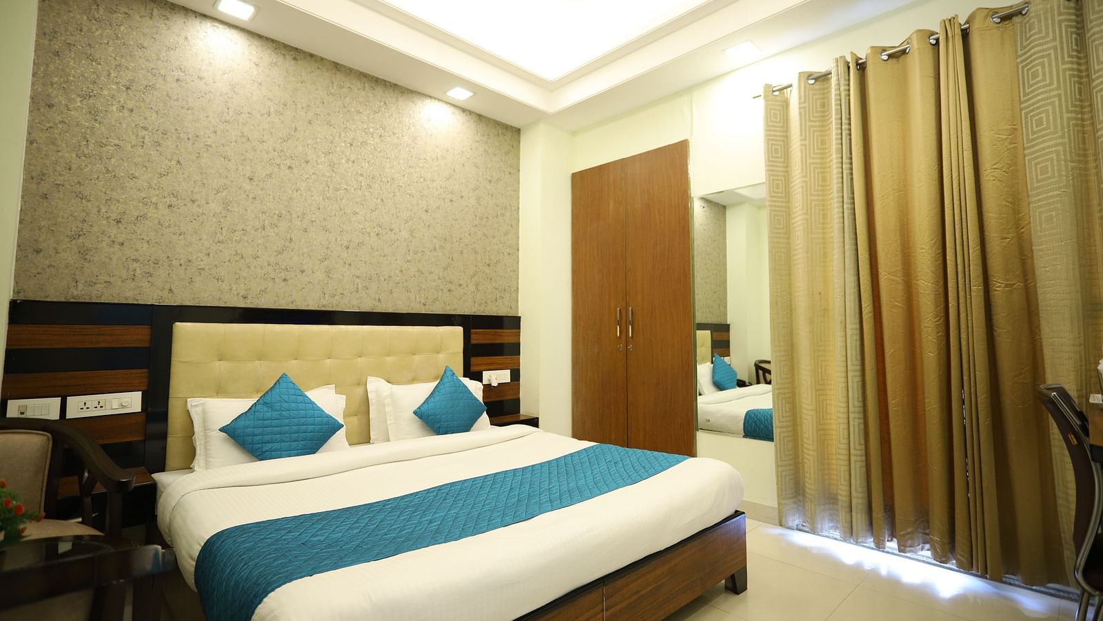 A well-lit room includes a double bed with blue accents, wooden furniture, and a mirror on the wall - Asian Suites Cyber Park, Gurgaon