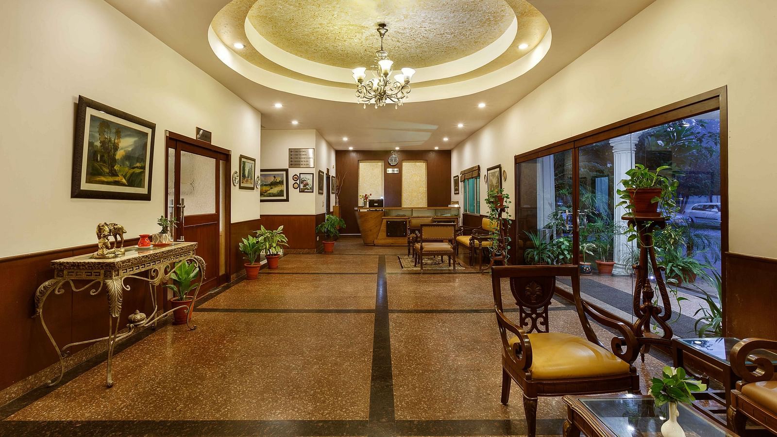 A corridor adorned with decorative frames, featuring high tables topped with flower vases and indoor plants, along with a few chairs arranged along the sides