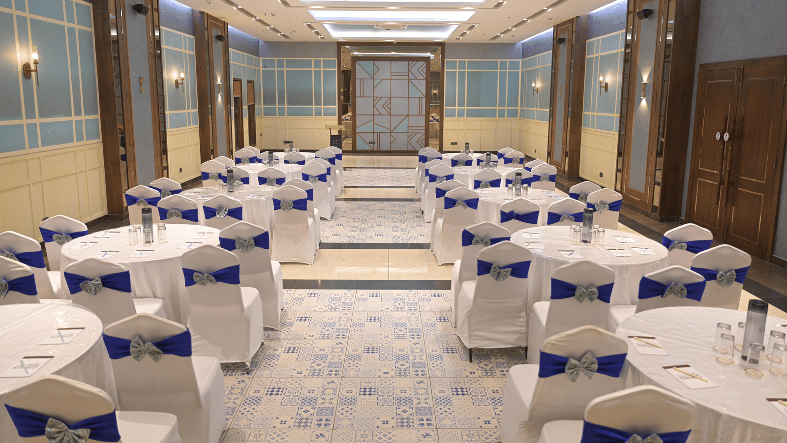 A straight, centred view of the banquet hall with round tables and blue-accented chairs at Hotel Sonar Bangla Mayapur.