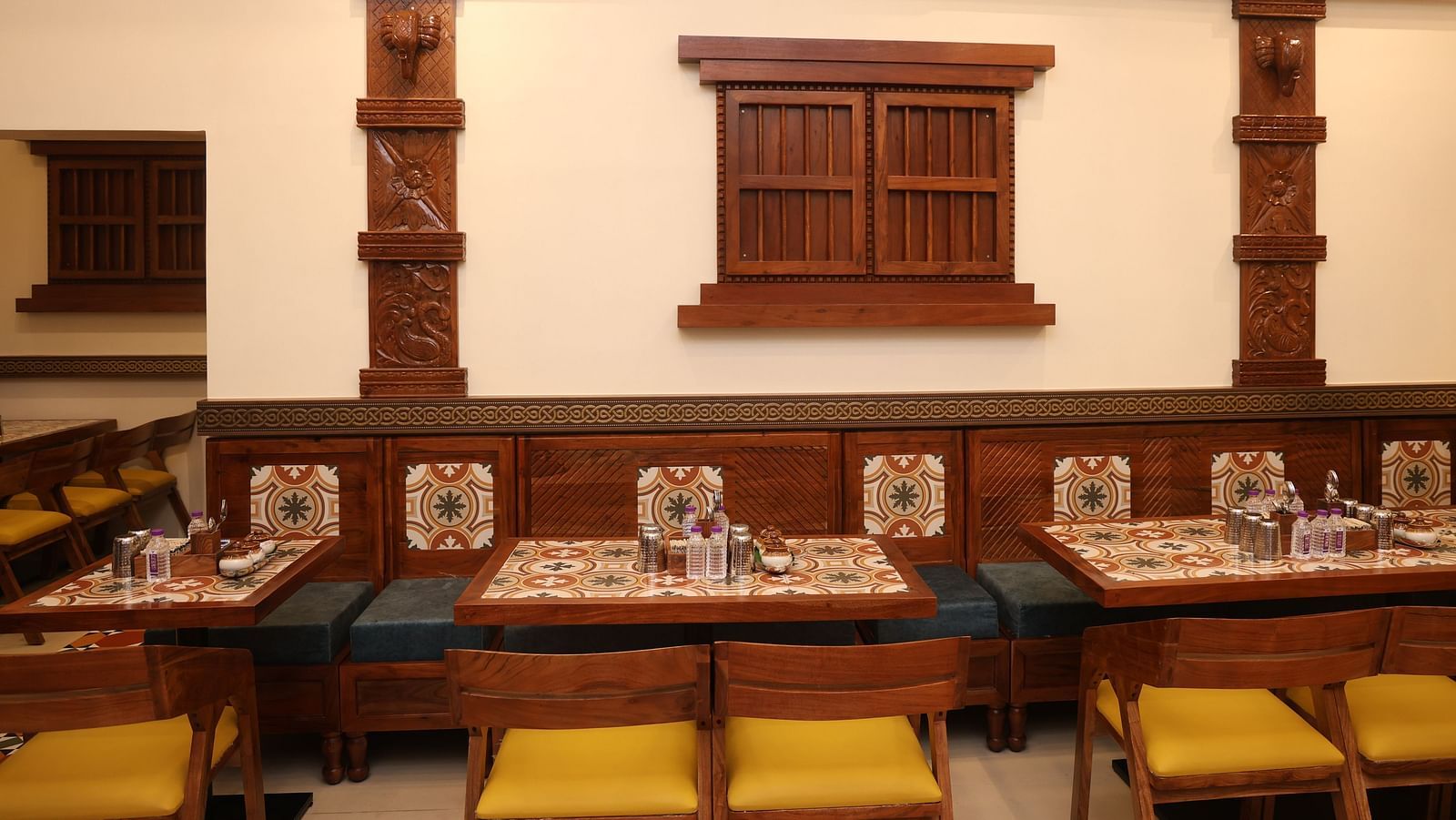 Indoor dining setup with traditional wooden décor, intricately carved pillars, tiled table settings, and cushioned wooden chairs with yellow seats in a warmly lit restaurant.