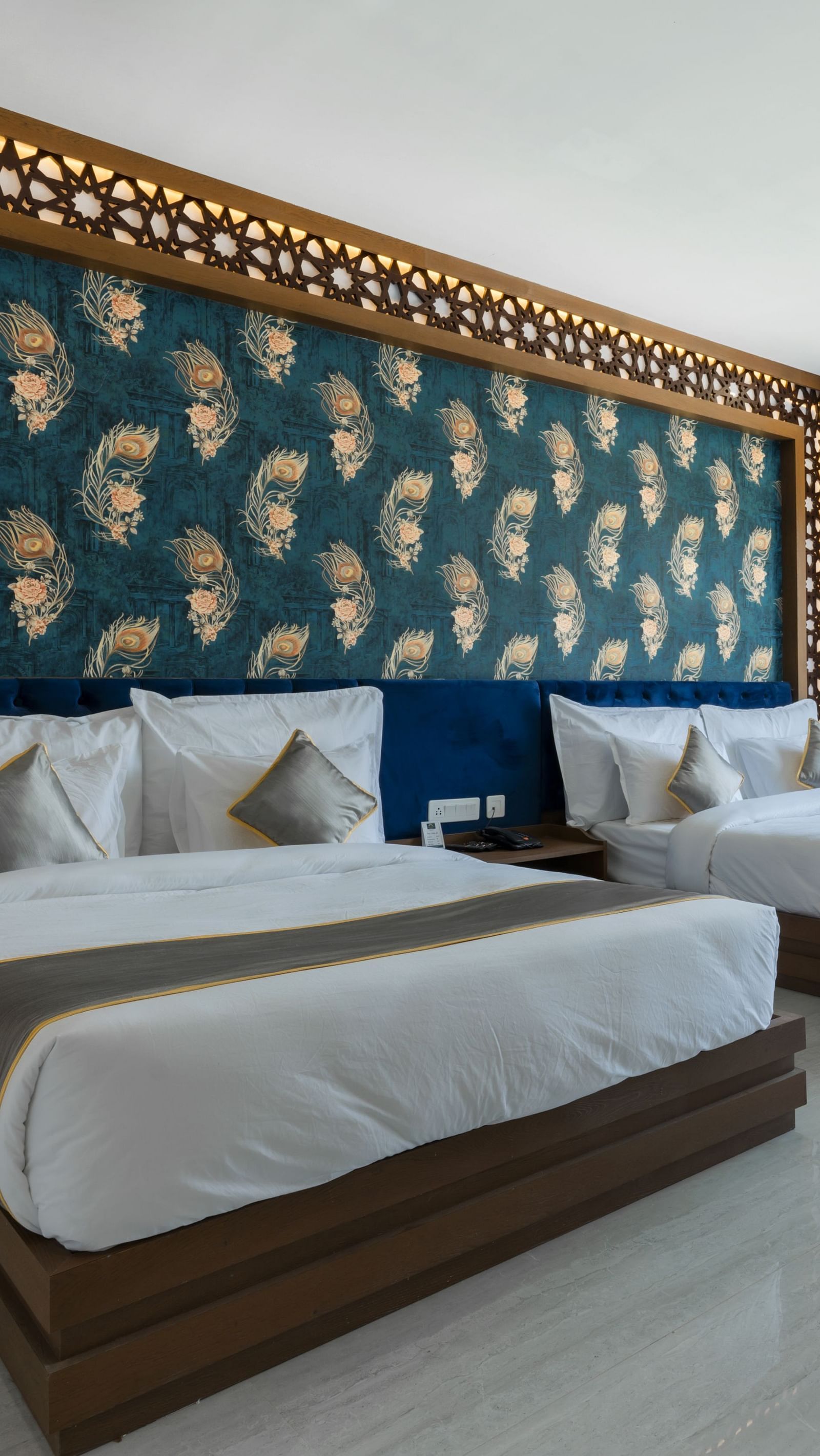 A bedroom at the Bamboo Saa Mulberry Resort featuring two twin beds with white linens and grey throws. A decorative wall panel with a blue and gold pattern is above the beds and a large windows is visible.