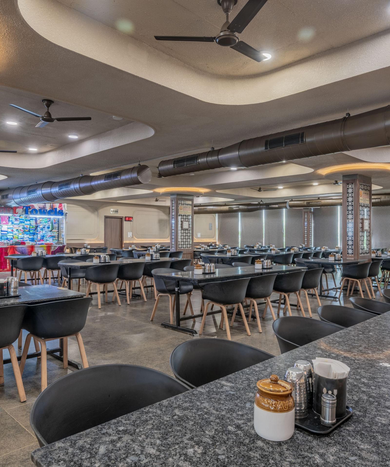 A restaurant interior with sleek tables, ambient ceiling lights, and neatly arranged seating at Hotel Highway King, Neelka.
