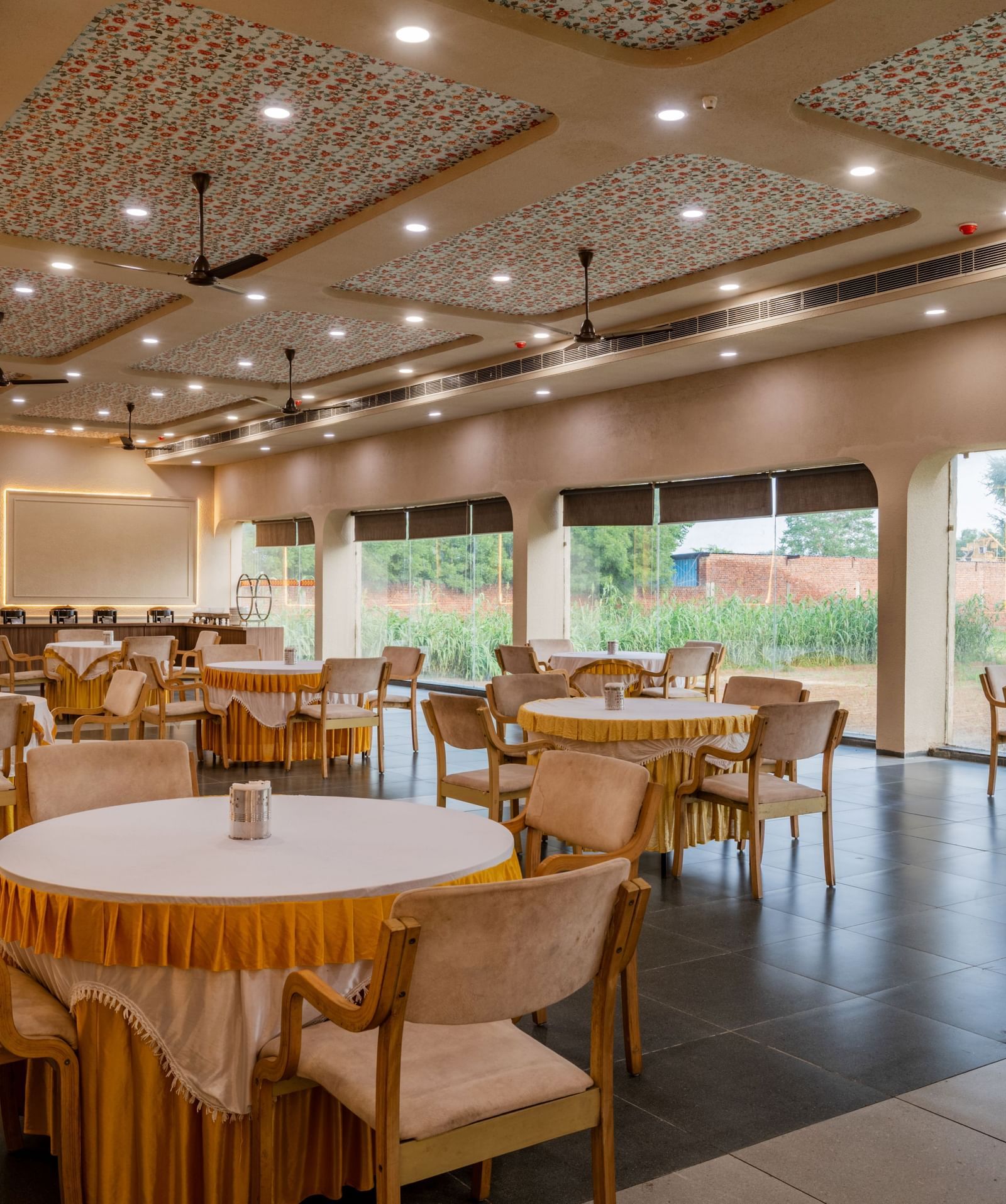 Spacious banquet area with large windows and round tables surrounded by chairs at Hotel Highway King, Neemrana. 