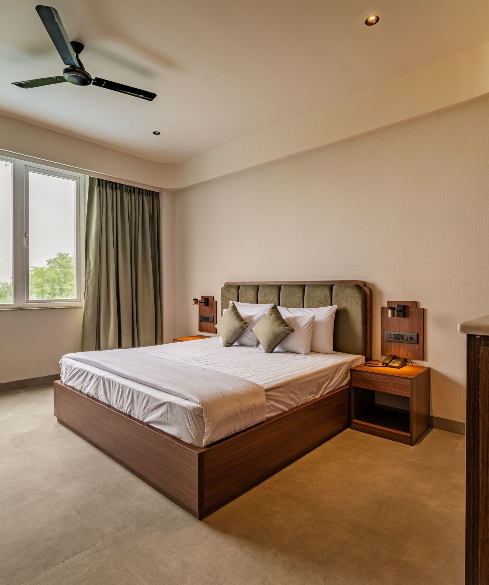 Diagonal view of a spacious room with a large bed, bedside tables, wide windows with curtains to the sides, and a wooden table with a plant at Hotel Highway King, Neemrana.
