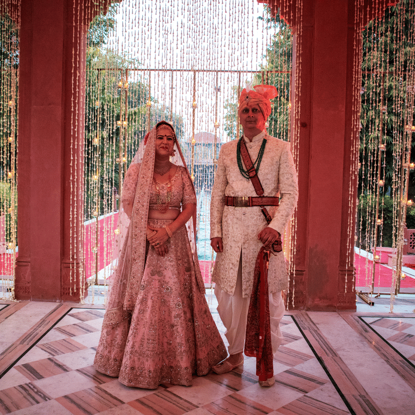 A bride and groom stand together in traditional outfits, framed by a curtain of hanging flowers- Umaid Palace - Best Luxury Heritage Resort Near Jaipur. 