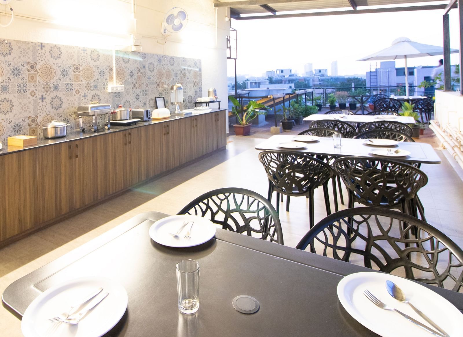 A dining space with tables and chairs under a partial roof, featuring a buffet setup along one side at Sanctum Suites Indiranagar, Bangalore.