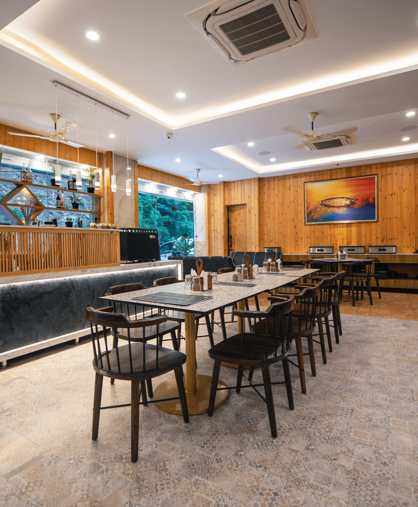 A restaurant at The Tattva Devaprayag with a long wooden table with black chairs and a bar.