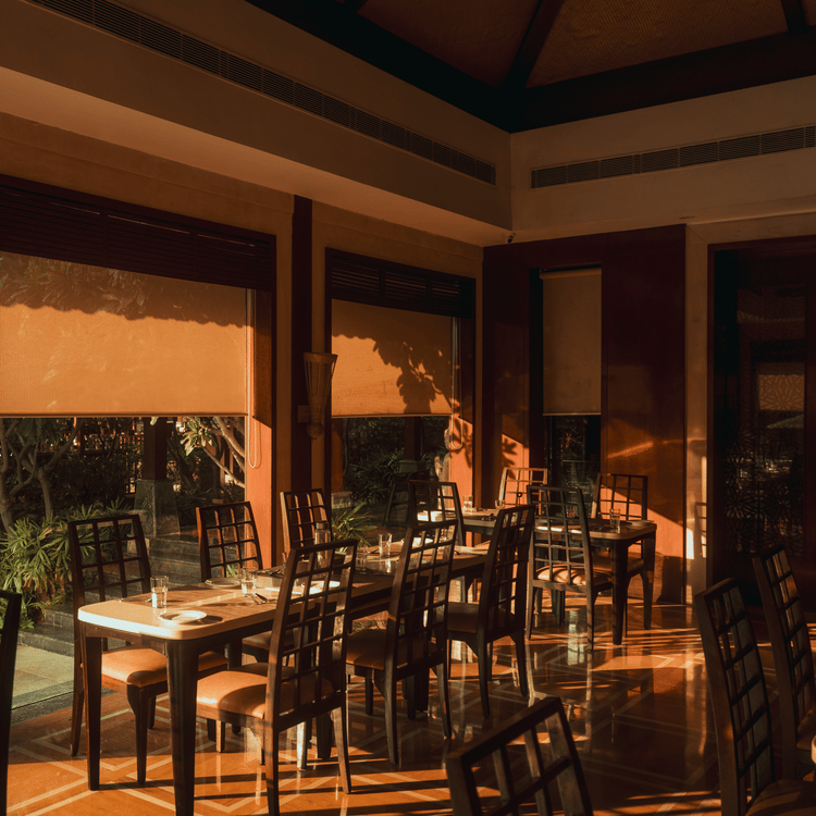 A well-lit dining area at The Ananta Udaipur, featuring wooden tables and chairs, large windows, and a view of the lush greenery.