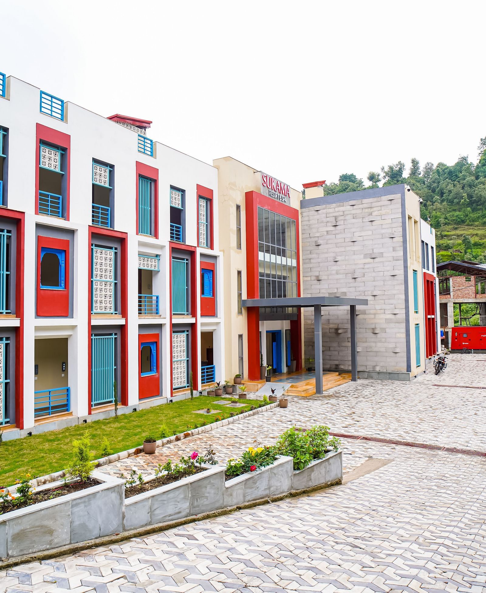 The exterior of a modern building with red and white sections and a paved courtyard is visible | Sukama Resort & Spa, Nainital